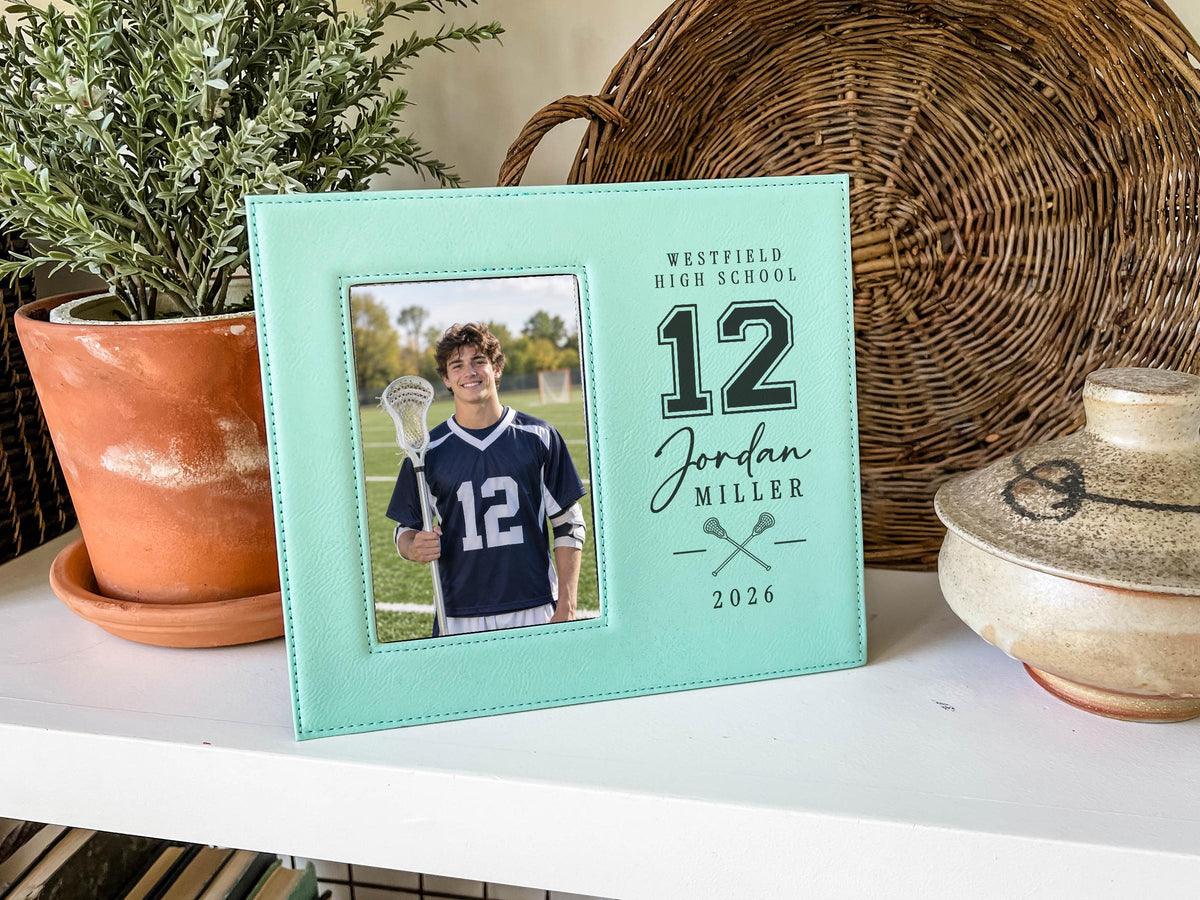 Teal Varsity Lacrosse frame mockup beside a potted plant and woven tray, showing a lacrosse player portrait and personalized number 12, Jordan Miller and 2026 engraving.