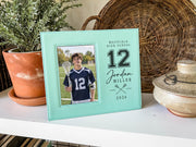Teal Varsity Lacrosse frame mockup beside a potted plant and woven tray, showing a lacrosse player portrait and personalized number 12, Jordan Miller and 2026 engraving.