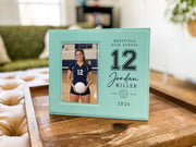Edited teal Varsity Volleyball frame mockup on a wooden tray, with an illustrative volleyball player portrait, varsity number 12, Jordan Miller, school name and year.