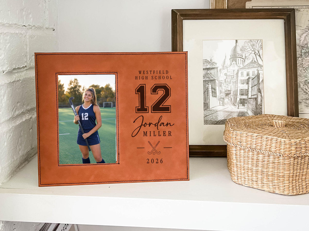 Warm reddish-brown Varsity Field Hockey frame on a white shelf, with an illustrative player portrait, number 12, Jordan Miller, crossed field hockey sticks and 2026.