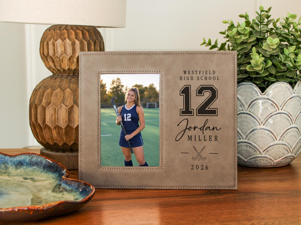 Varsity Field Hockey frame beside a lamp and plant, showing an illustrative player portrait and personalized school, number 12, Jordan Miller and 2026 engraving.