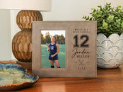 Varsity Field Hockey frame beside a lamp and plant, showing an illustrative player portrait and personalized school, number 12, Jordan Miller and 2026 engraving.