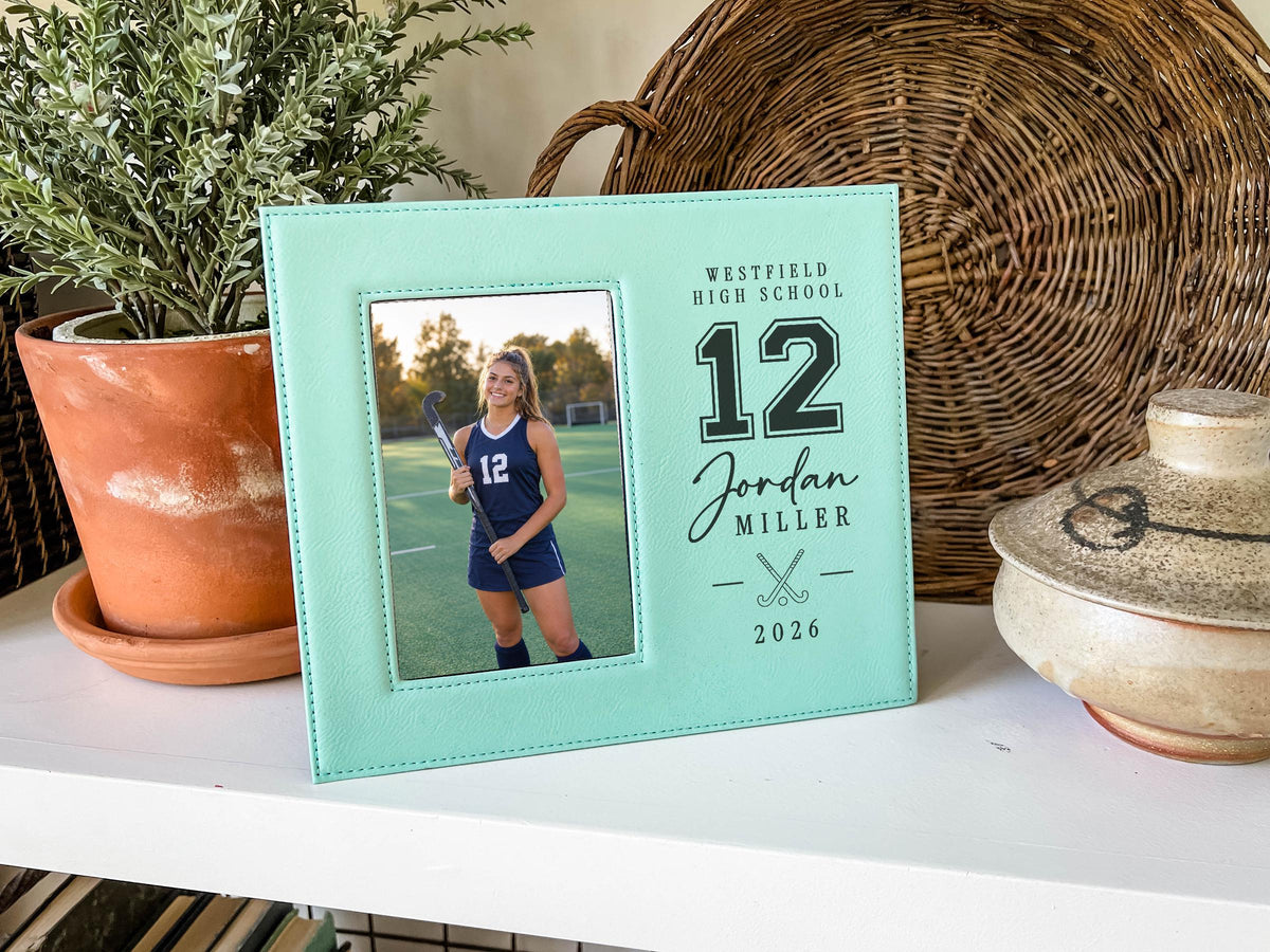 Teal Varsity Field Hockey frame beside a potted plant and woven basket, with an illustrative player portrait and sample school, name, number and year engraving.