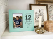 Teal Varsity Football frame mockup on a shelf beside a candle, with a sample player portrait, large jersey number 12, school, name, football illustration, and year.