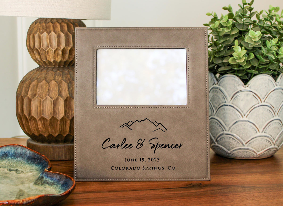 a picture frame with a mountain design, placed on a wooden surface. The frame has a blank photo inside and a personalized text that reads "Carlee & Spencer JUNE 19, 2023 COLORADO SPRINGS, CO". There is also a potted plant and a bowl in the background.