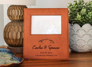 a wooden picture frame with a mountain design, placed on a wooden surface next to a potted plant and a decorative vase.