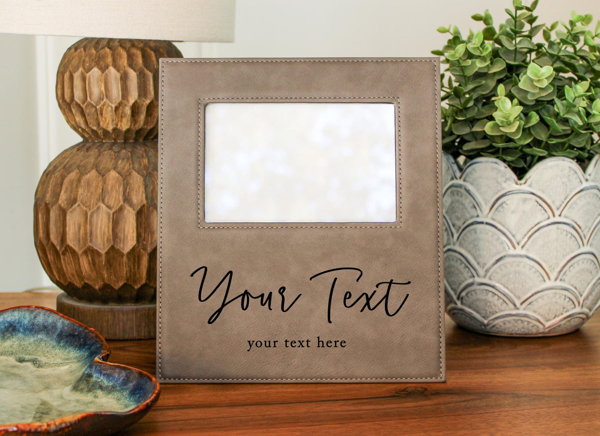 A gray picture frame with a blank space in the center, placed on a wooden surface. The frame has a personalized text on it, and there are decorative items such as a potted plant and a ceramic bowl in the background.