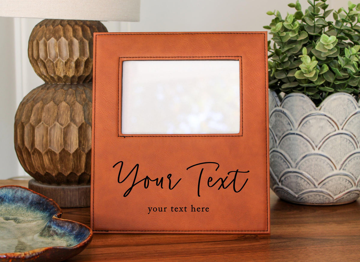 a brown leather picture frame with a blank white space in the center, placed on a wooden surface. The frame has a personalized message that reads "Your Text" and "your text here". In the background, there are decorative items such as a lamp and a potted plant.