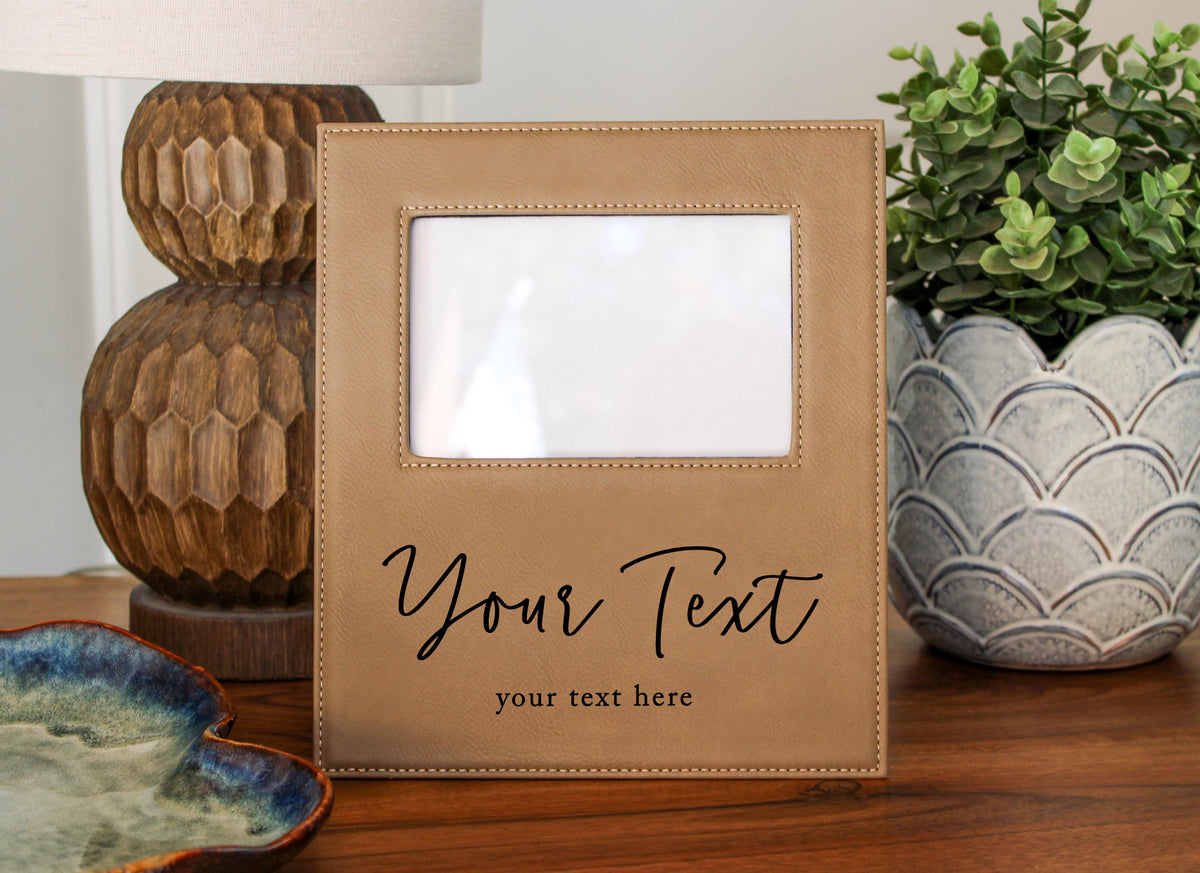 a tan picture frame with a blank photo space in the center, placed on a wooden surface. The frame has a decorative design on the sides. In the background, there are two potted plants, one on the left and one on the right, as well as a lamp and a ceramic bowl. The text "Your Text" is visible on the frame, suggesting that it is a customizable photo frame.
