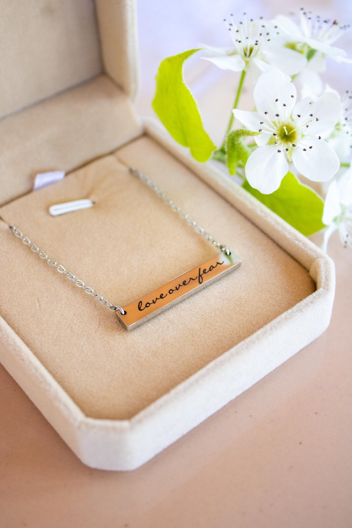 A silver bar necklace engraved with 'love over fear' in black script, presented in a beige velvet jewelry box with white flowers in the background.