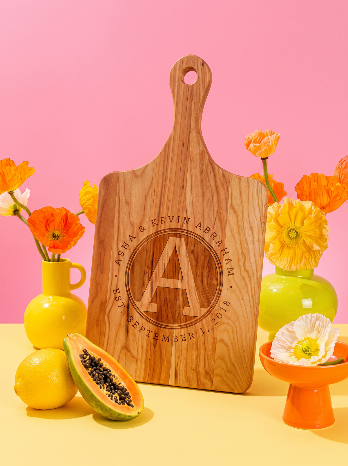 Wood paddle cutting board with a circular initial monogram, names and date engraving, styled with colorful flowers, view 1.
