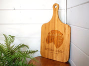 A wooden cutting board with a custom logo engraving, placed against a white wall next to a green plant.